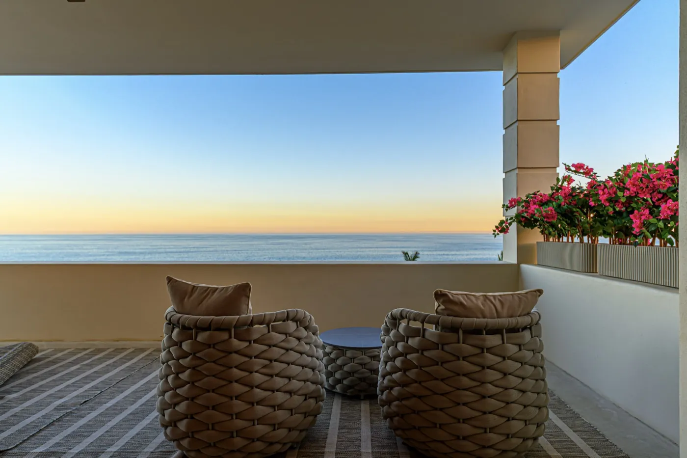 Covered terrace with woven armchairs framing a calm sea view and bougainvillea at dusk.