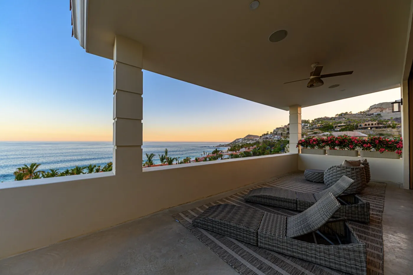 Covered terrace with woven loungers framing a tranquil sea and coastline under a soft sunset sky.