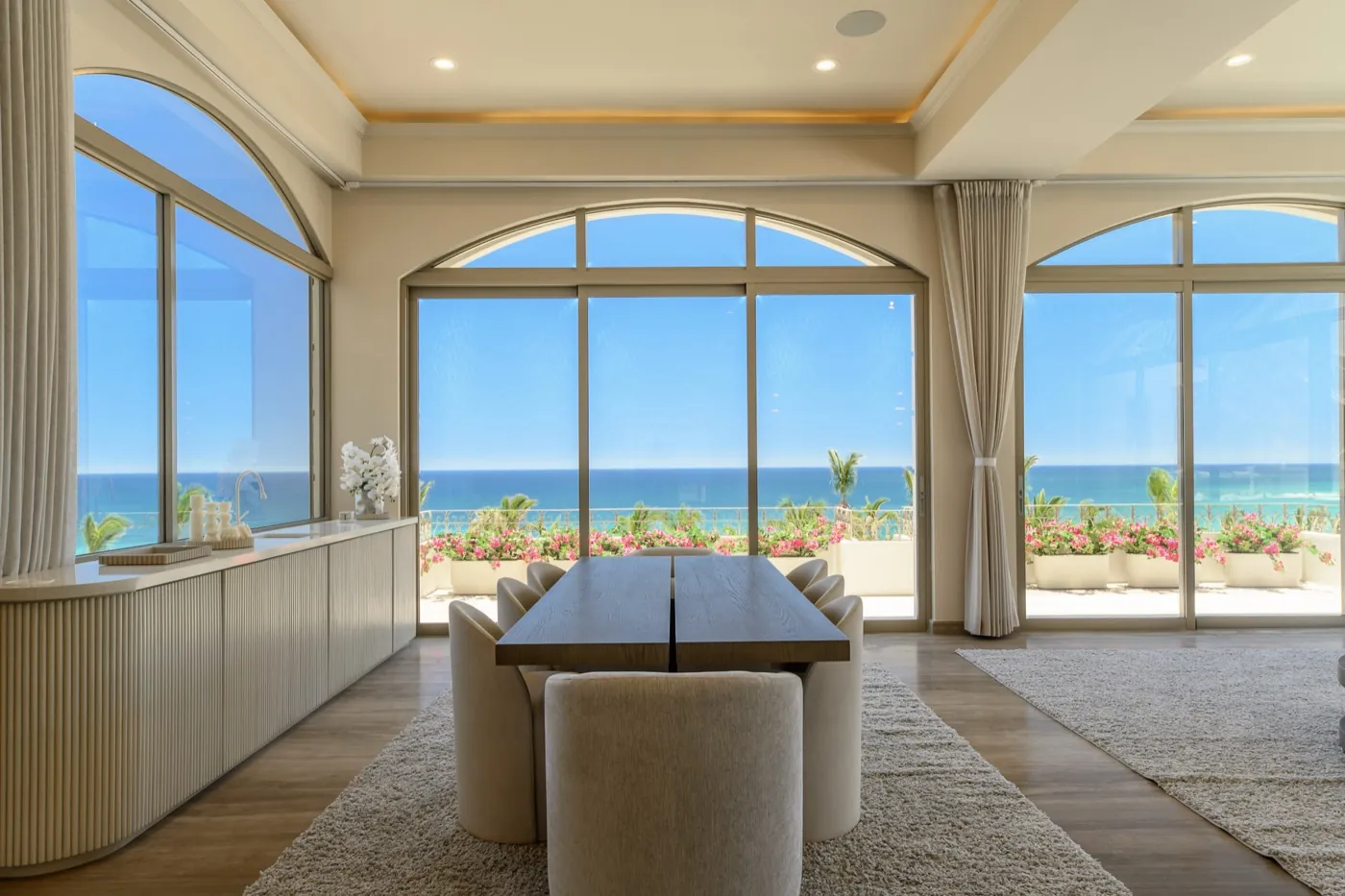 Dining room framed by arched windows, the table set before a bougainvillea terrace and ocean.