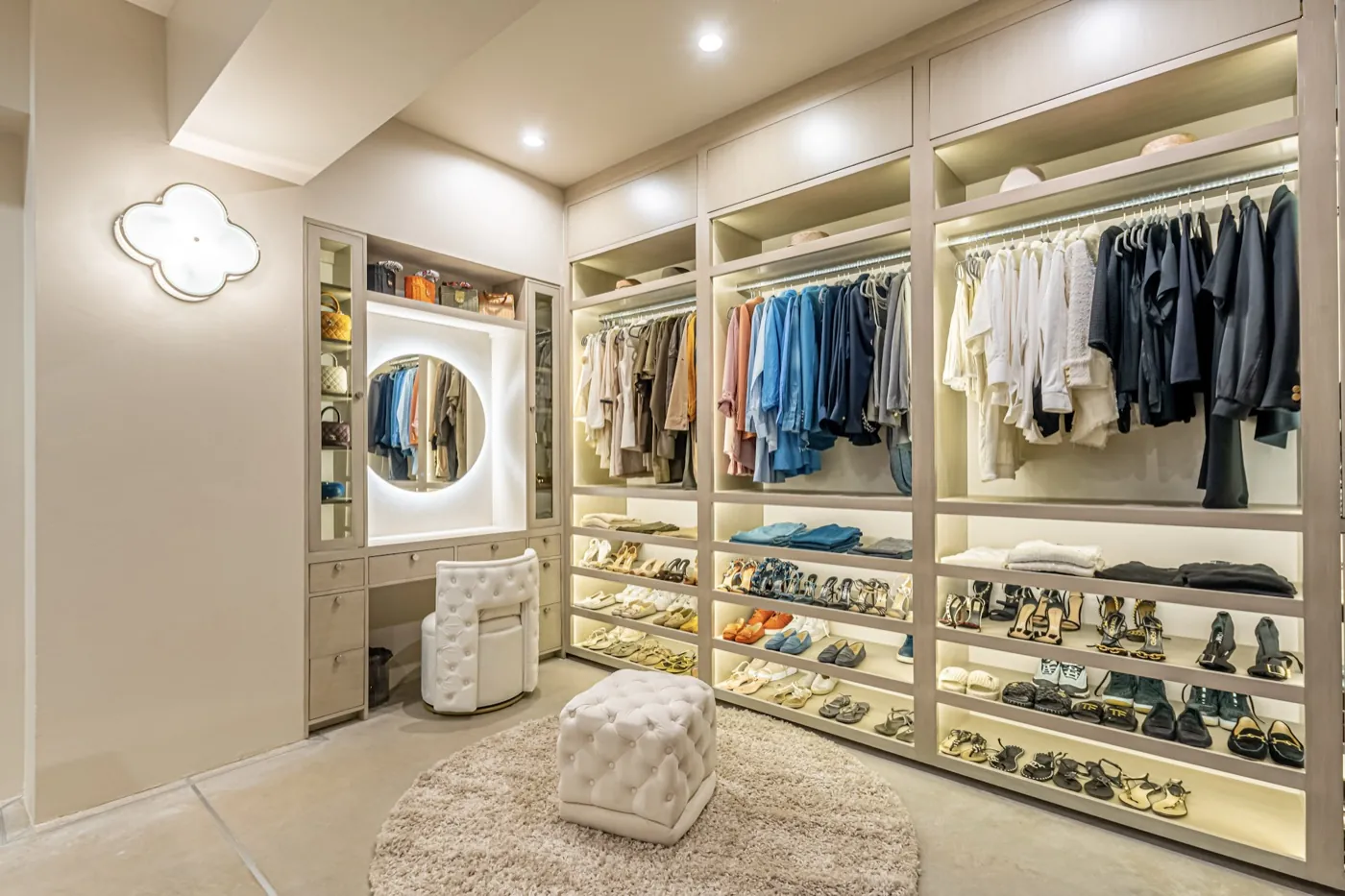 Dressing room with backlit open shelving, an illuminated round mirror and a tufted ottoman on a soft rug.