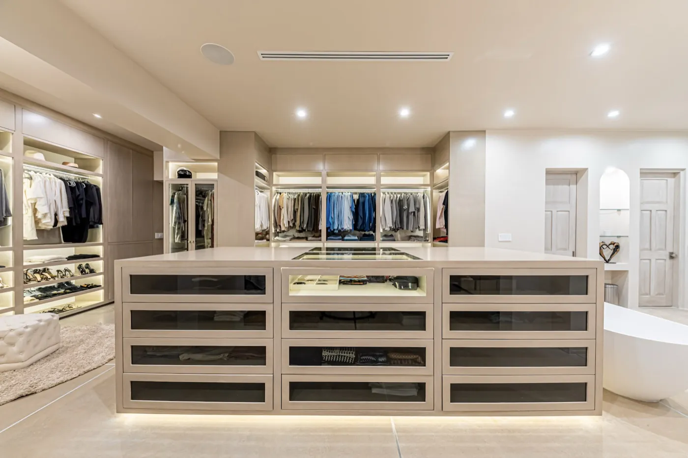 Spacious dressing room with a glass-fronted central island, backlit wardrobes and a freestanding bath beyond.