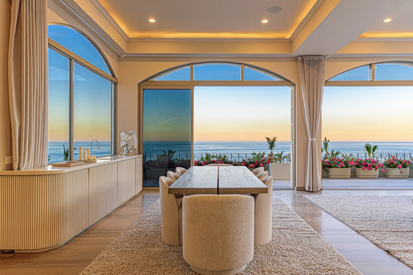 Formal dining room with arched windows opening to a flowering terrace and the sea at sunset.