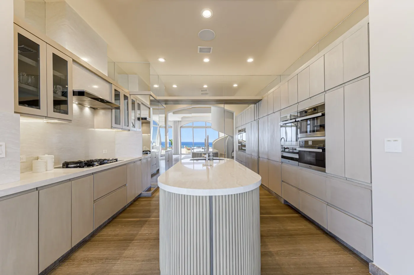 Galley kitchen in pale oak with a fluted marble island and a distant sea view framed by glazing.