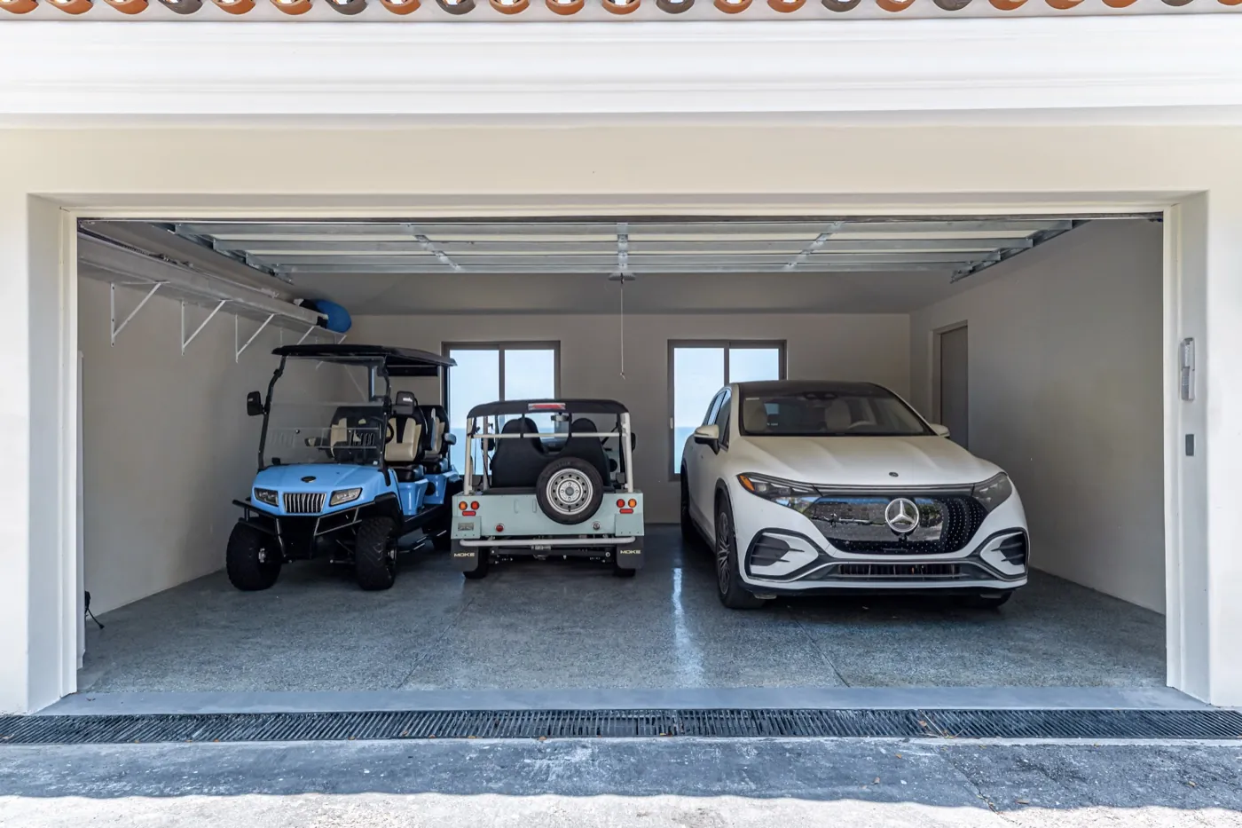 Two-bay garage holding a white Mercedes, a vintage open-top jeep and a blue golf cart on polished concrete.