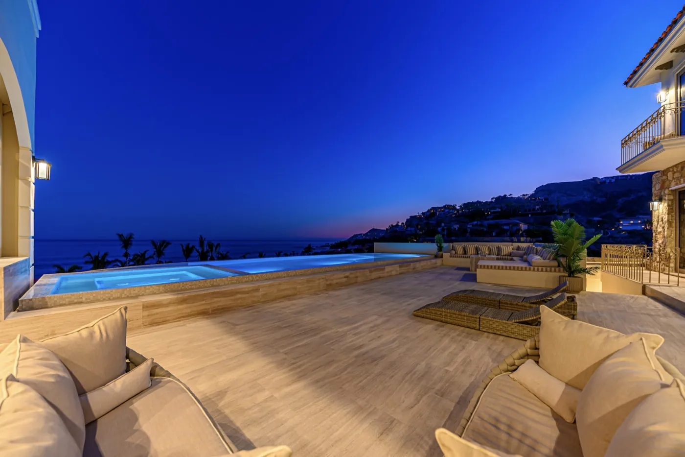 Dusk terrace with cushioned seating beside an infinity pool overlooking the sea under a deep blue sky.