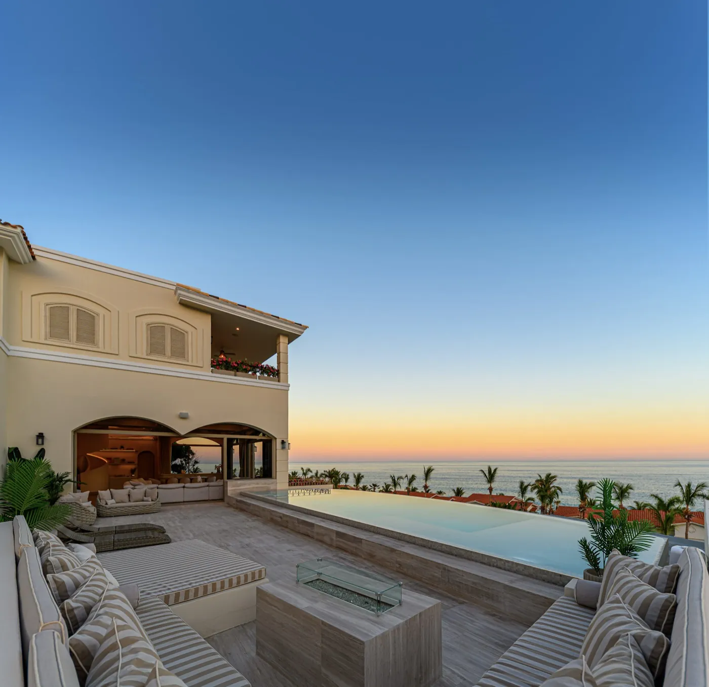 Infinity pool meeting the horizon at twilight, framed by striped daybeds and a glass-edged fire table.