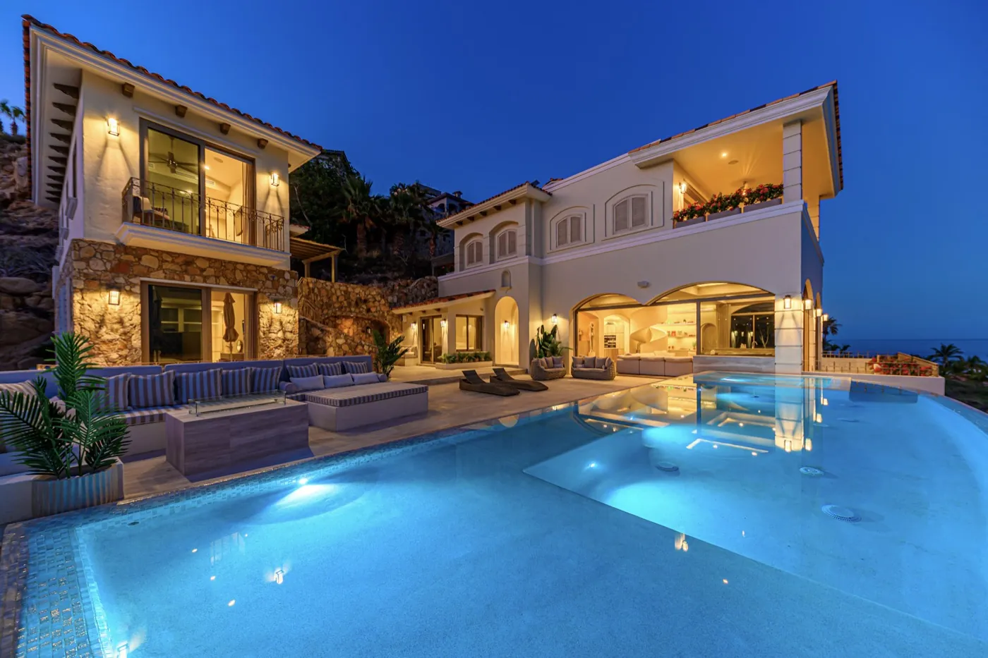 Illuminated infinity pool before the stone-and-stucco villa glowing against a deep blue dusk sky.