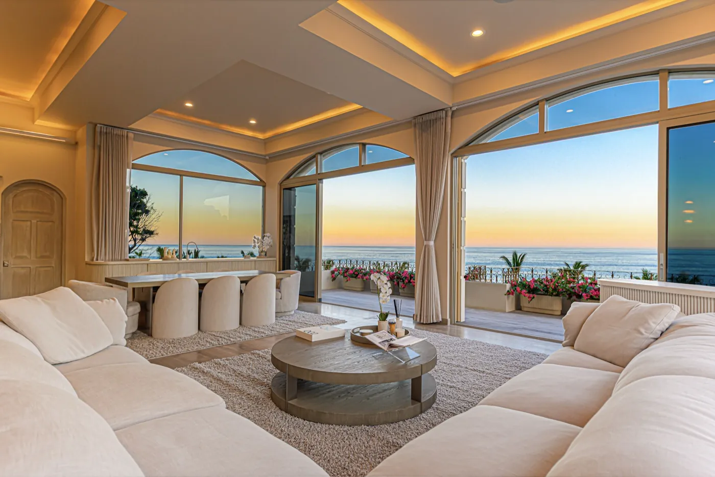 Open living and dining room with plush sofas opening to an ocean terrace at sunset.