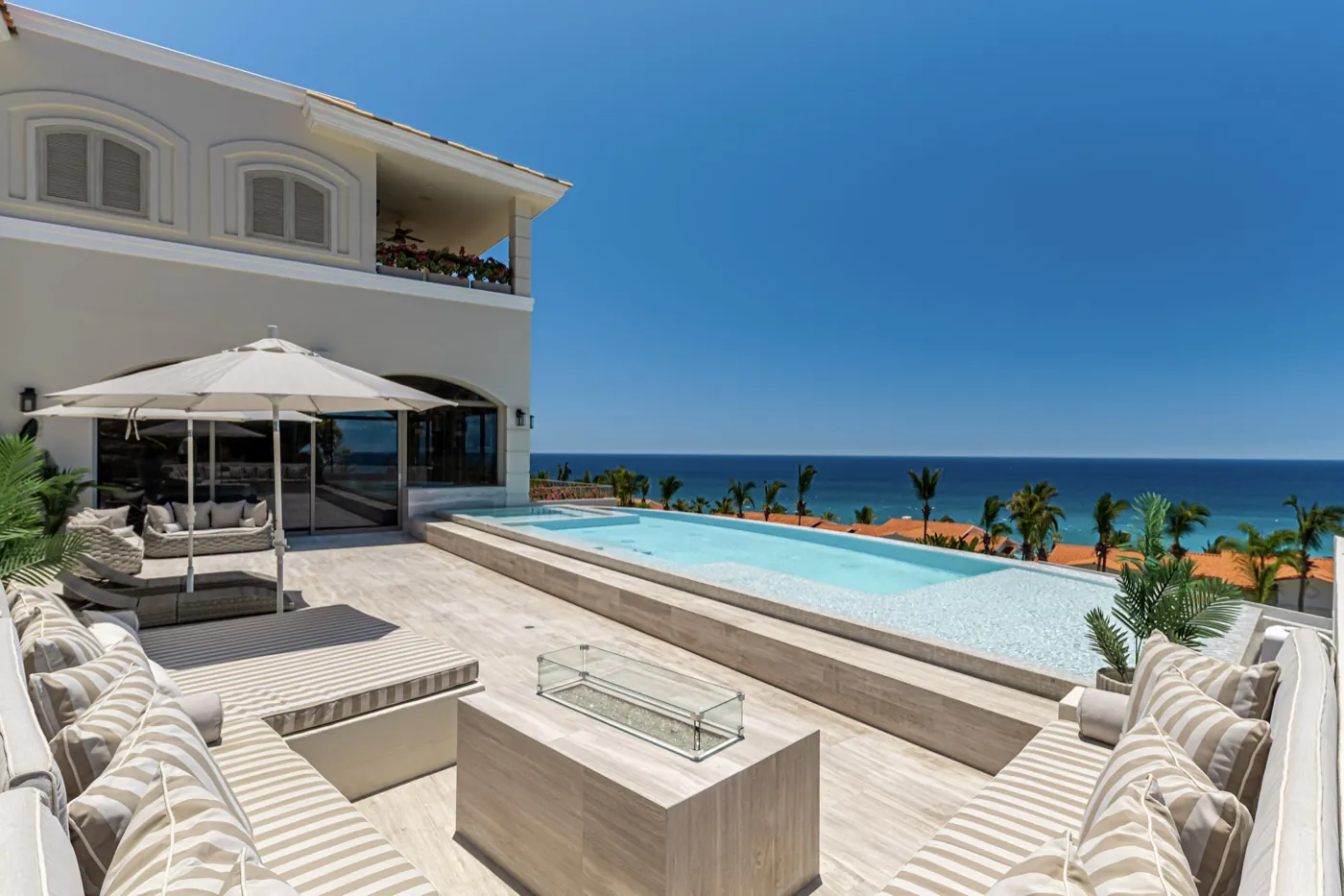 Sun-bleached stone terrace with cushioned loungers, a parasol and a fire table beside the infinity pool and sea.