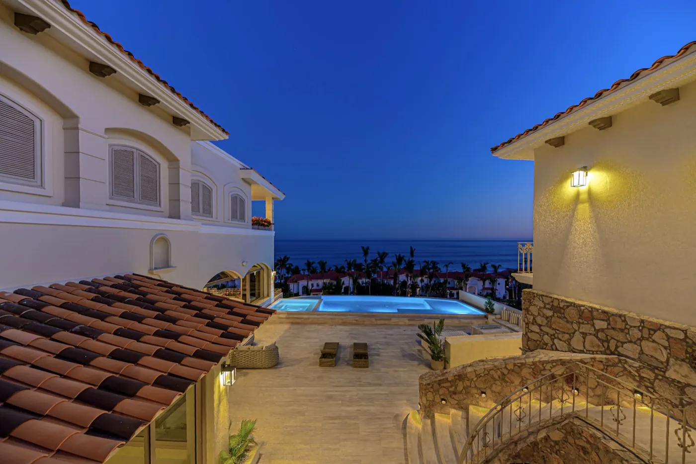 Stone courtyard and lit pool between villa wings under a deep blue twilight sky above the sea.