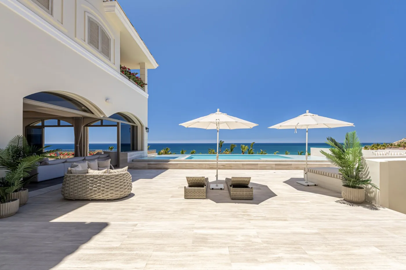 Sunlit travertine pool terrace with twin cream parasols, woven seating and a calm sea beyond.