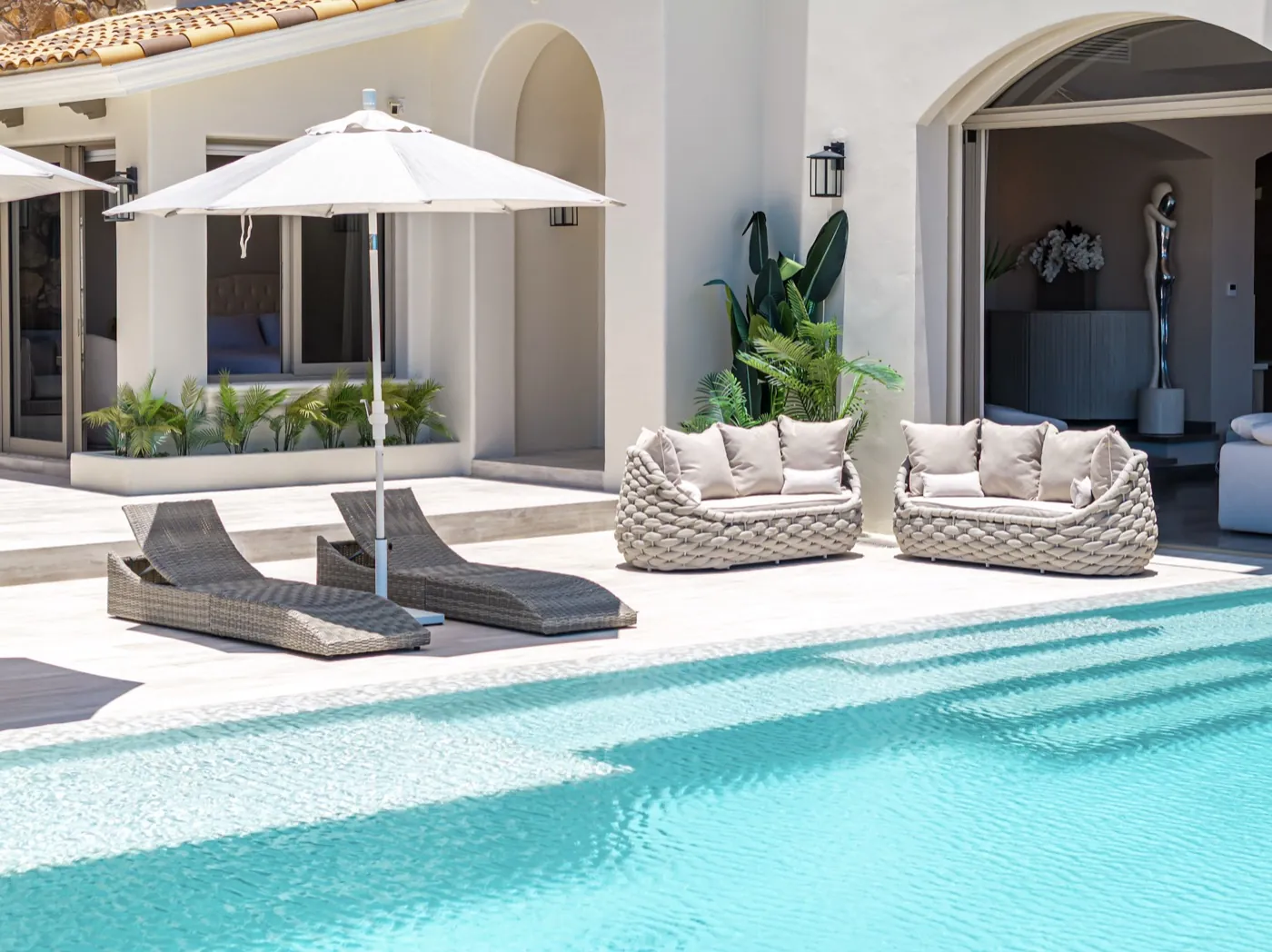 Poolside terrace with woven loungers, a sofa and parasol beside a turquoise pool and arched villa.