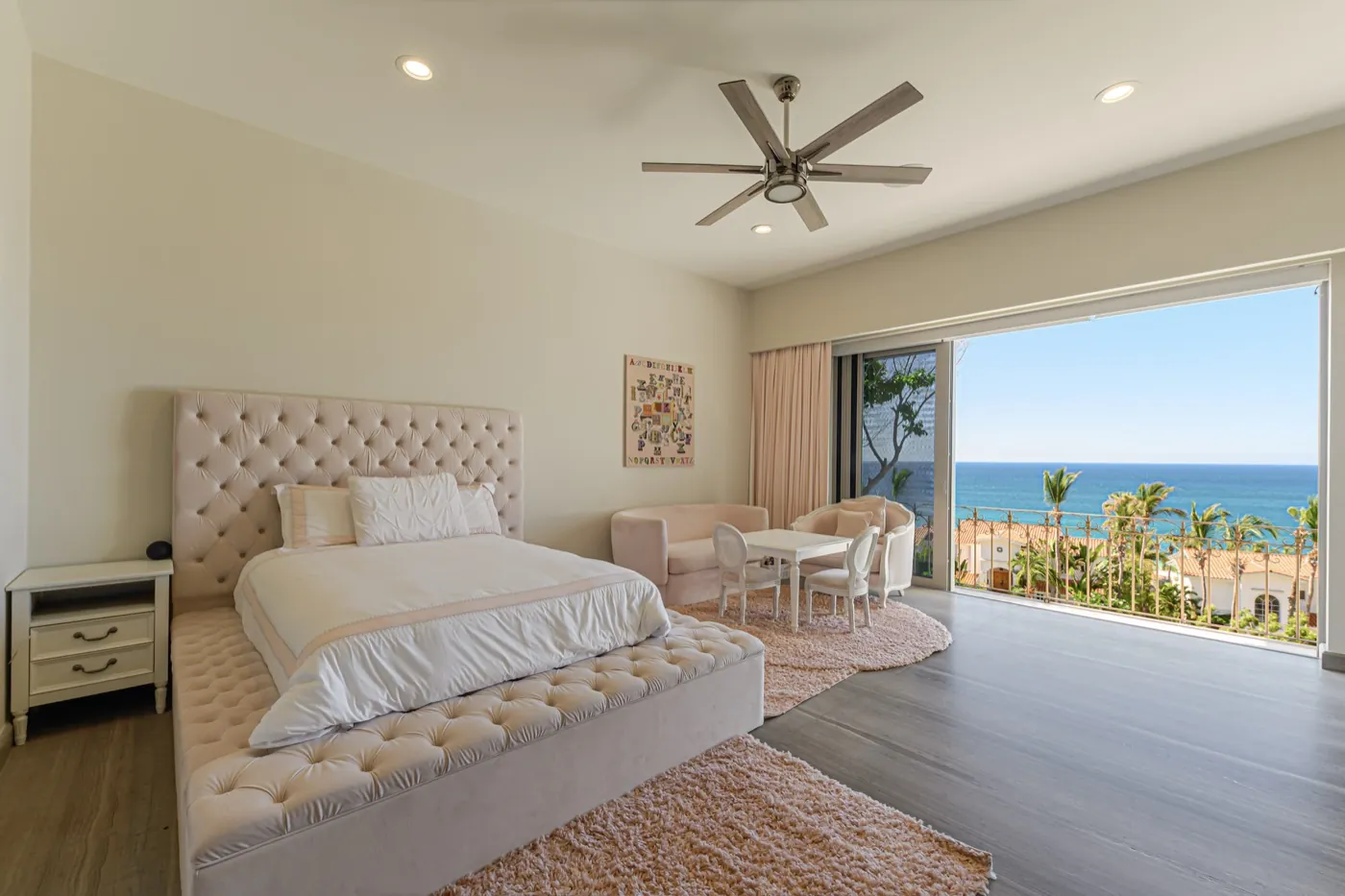 Soft blush primary bedroom with tufted bed opening through sliding glass to a palm-fringed ocean view.