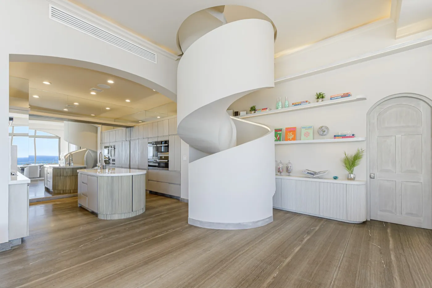 Open-plan living space where a sculptural white spiral staircase rises beside a curved limestone kitchen island.