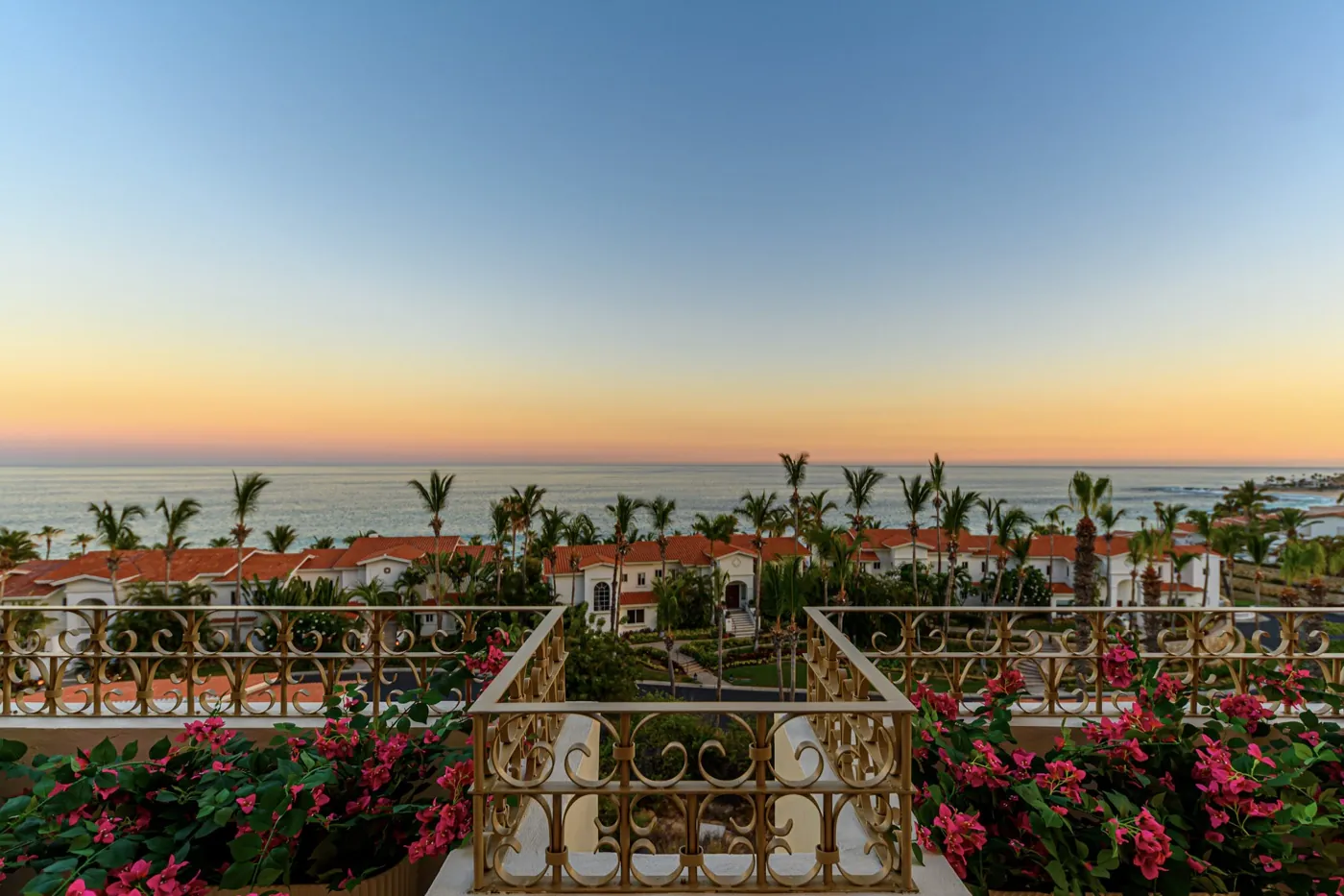 View from an ornate gilt balustrade over palms and tiled rooftops to the sea at dusk.