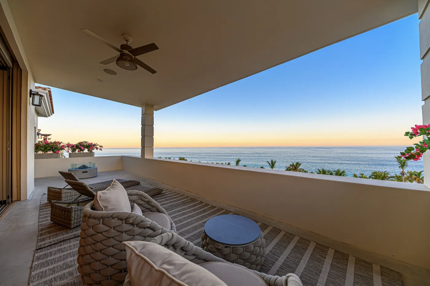 Covered ocean terrace with woven loungers and a ceiling fan at golden sunset over calm water.