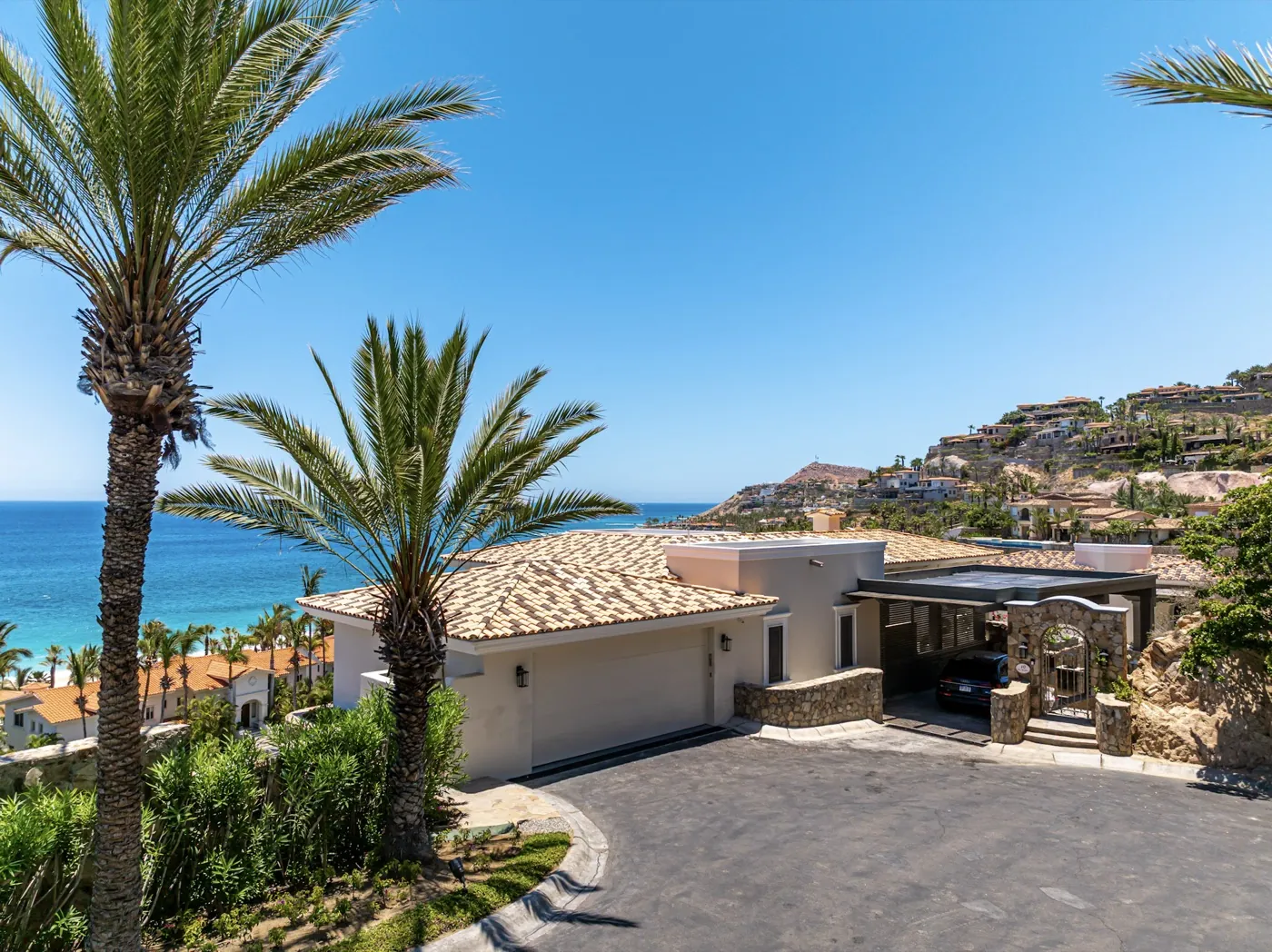 Villa entrance and tiled-roof garage framed by tall palms above a curving driveway and the distant ocean.