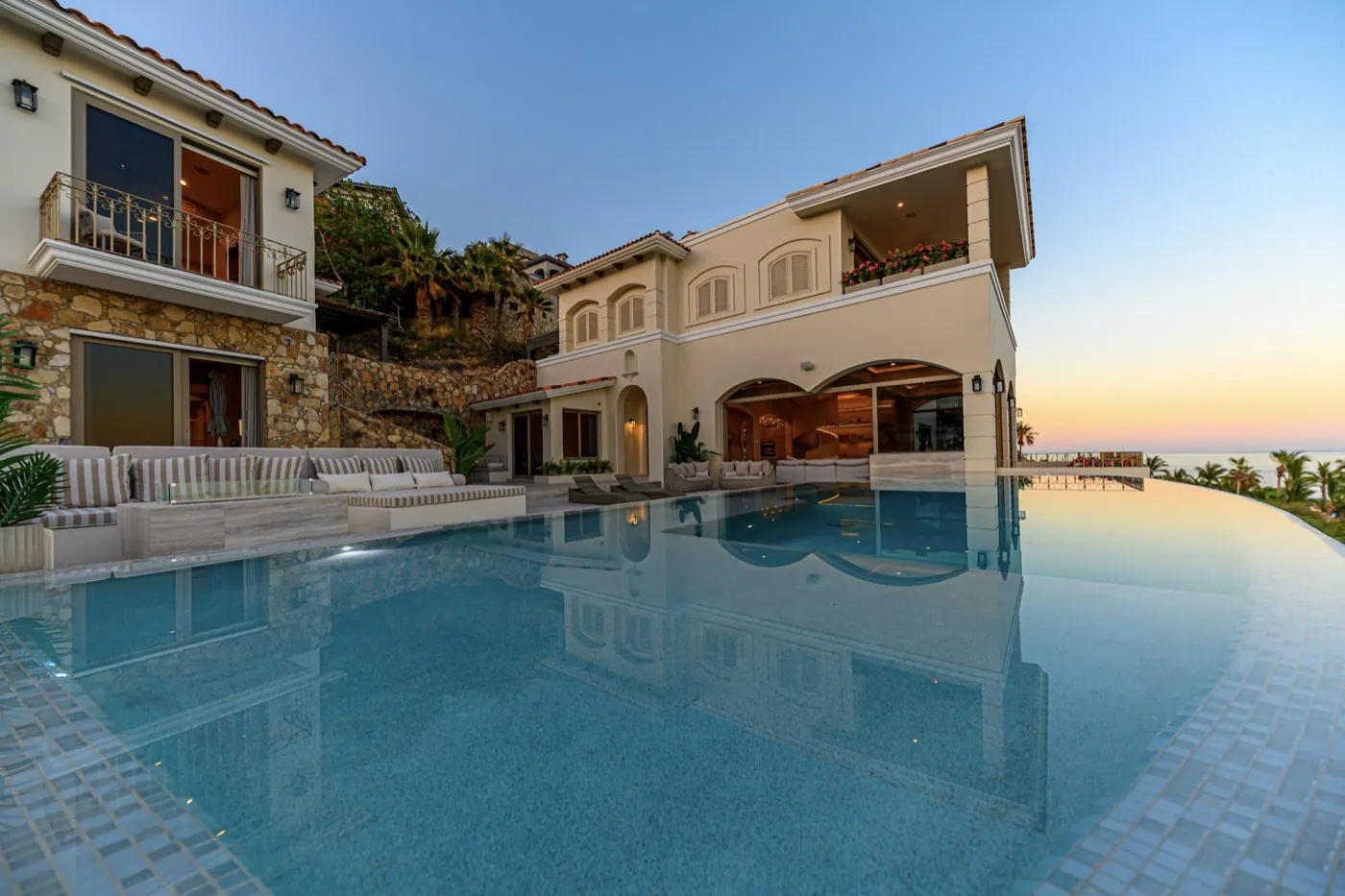 Stone-and-stucco villa facade mirrored in a calm infinity pool at dusk above the sea.