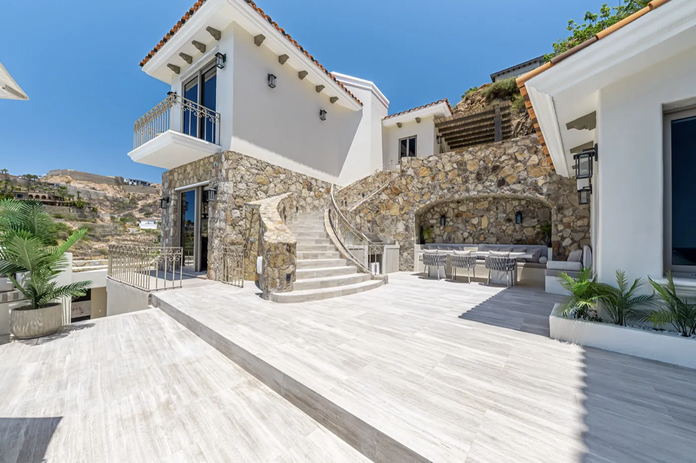 Travertine courtyard with a curved stone staircase rising between natural-stone walls under clear skies.